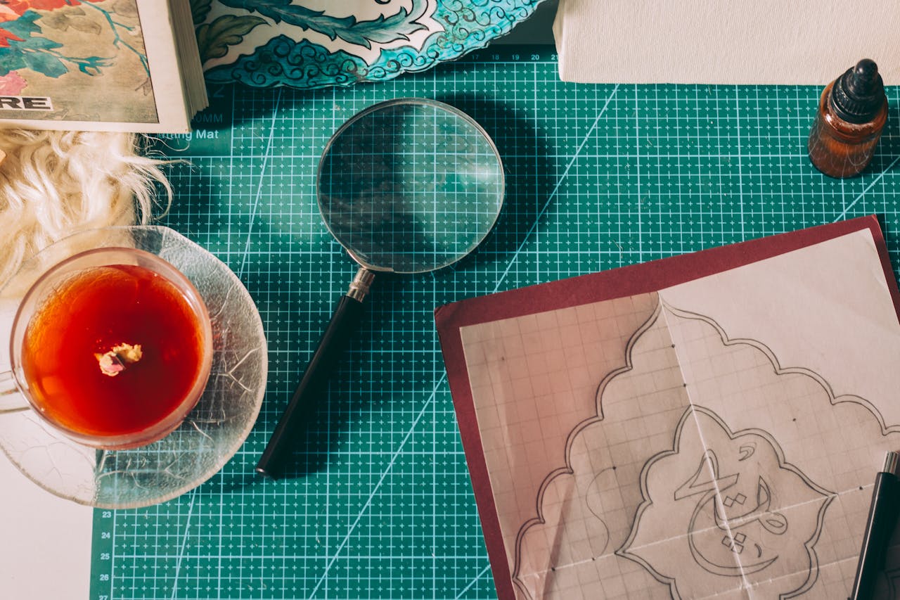 A cozy workspace with art supplies, tea, and a magnifying glass on a cutting mat.