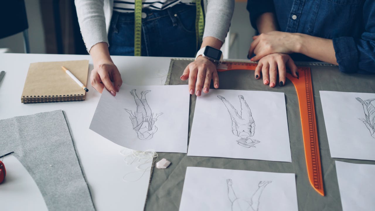Two designers discuss fashion sketches and fabrics at a workspace.